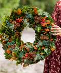 Person holding a festive wreath with green leaves, pinecones, and berries against a blurred natural background.
